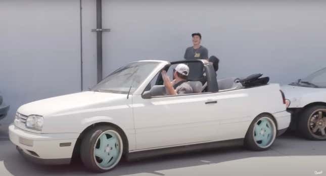 Two men attempt to park a white VW Cabrio in front of a gray wall, unsuccessfully.